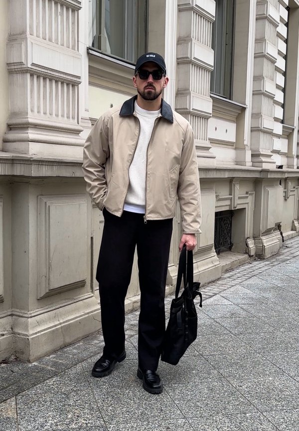 Moški, oblečen v bež jakno, belo jopico, črne hlače, črno kapo in sončna očala, nosi črno torbo čez ramo na mestnem pločniku.