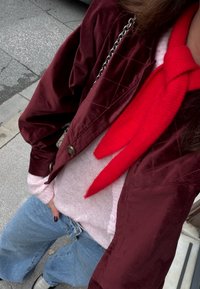 Personne portant une veste matelassée bordeaux, un pull rose pâle, un jean bleu et une écharpe rouge vif, debout sur un trottoir.