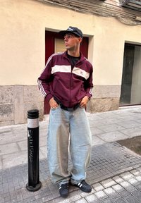 Jachetă de trening vișinie cu dungi albe, tricou negru, blugi lejeri de culoare albastră deschisă și adidași negri. Stând aproape de un stâlp de stradă negru.