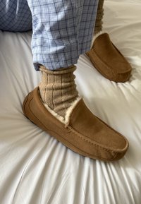 Bruna mockasin med pälsfoder, rundad tå och tonal sömnad. Bärs med ribbade beige strumpor och rutig blåbyxor på vit sängklädsel.
