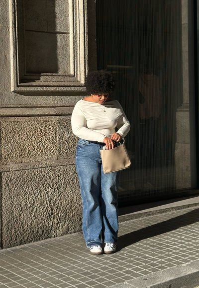 Blusa de manga larga beige claro, vaqueros azul holgados y zapatillas blancas. Sosteniendo un pequeño bolso beige. De pie contra una pared de piedra.