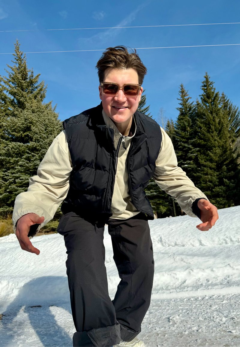 Junge Person mit Sonnenbrille, schwarzer Weste und beige Jacke hockt lächelnd im Freien auf Schnee, umgeben von immergrünen Bäumen und klarem blauem Himmel.