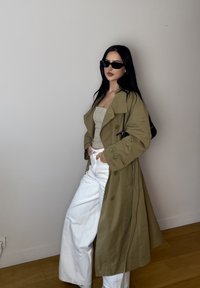 Larga gabardina verde oliva con solapas anchas, combinada con una blusa de cuadros clara y pantalones de pierna ancha blancos. Unas gafas de sol negras completan el look.
