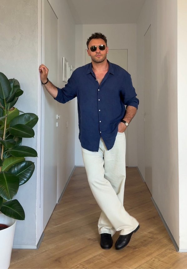 Homme portant des lunettes de soleil, une chemise bleu marine et un pantalon blanc, appuyé contre un mur blanc dans un couloir étroit avec un sol en bois et une plante verte.