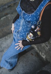 Jeansklänning med hängslen och mönster av halvmånar, kombinerad med en genomskinlig svart långärmad topp. Accessoarer inkluderar en färgglad nyckelringsleksak.