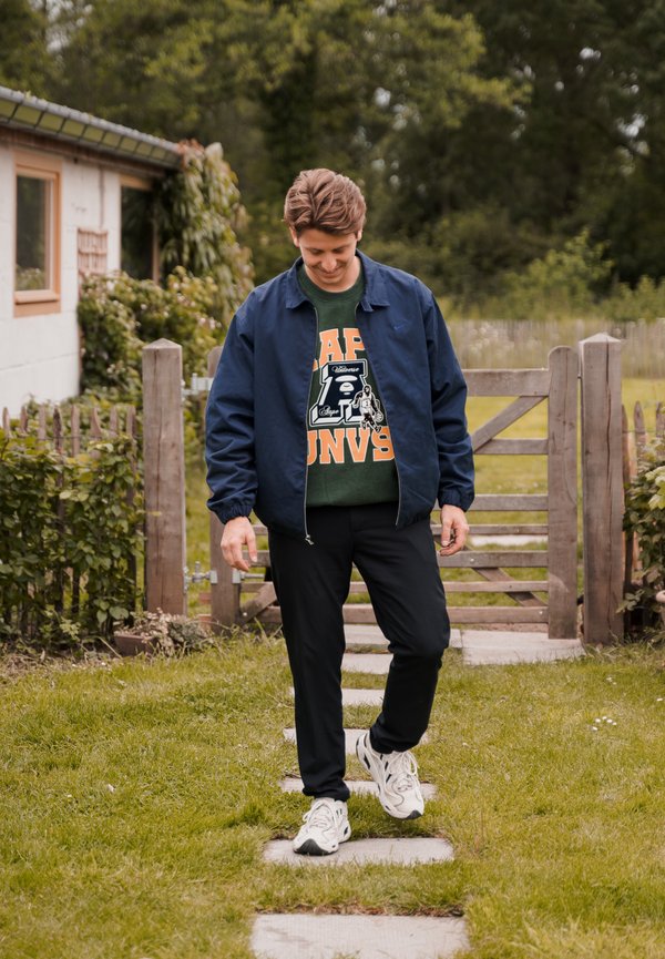 Conjunto informal que incluye una chaqueta azul marino, sudadera verde con texto naranja, pantalones negros y zapatillas blancas, caminando por un sendero de piedra en un jardín.