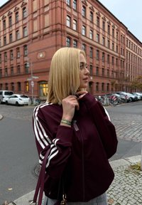 Vinrød lynlåstrøje med hvide striber på ærmerne, kombineret med grå ternede bukser. Tilbehør inkluderer et armbånd og en håndtaske. Baggrunden viser en bygning i røde mursten.