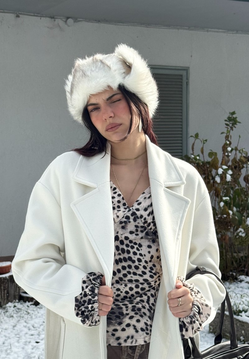 Weißer Wollmantel über einem schwarz-beigen, gepunkteten Oberteil, ergänzt durch einen Pelzhut. Strukturierte Ärmelbündchen und geschichteter Schmuck vervollständigen den Look.