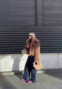 Femme portant une veste marron poilue, un jean large foncé, des chaussures roses, des lunettes de soleil, et tenant un sac à main beige tissé en marchant en extérieur.