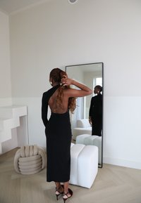 Schwarzes, figurbetontes Kleid mit einem langen Ärmel und offenem Rücken. Das Modell steht neben einem Spiegel und präsentiert das elegante Design und die schicke Silhouette.