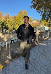 Άνδρας που στέκεται σε πλακόστρωτο πεζοδρόμιο κοντά σε κιγκλίδωμα, φοράει μαύρο μπουφάν, πουκάμισο με καρό σχέδιο, χακί παντελόνι, περιτριγυρισμένος από φθινοπωρινά δέντρα και καθαρό γαλάζιο ουρανό.