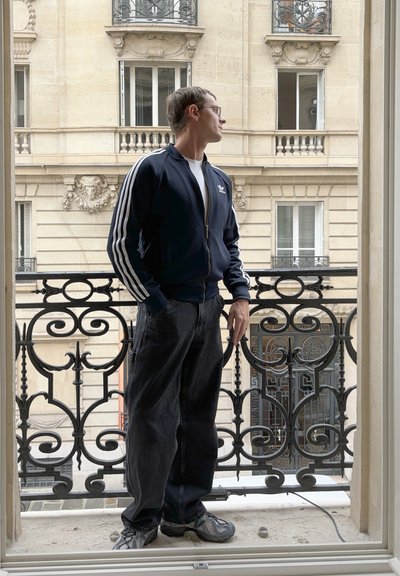 Chaqueta deportiva de Adidas con cremallera en color azul marino y rayas blancas, combinada con pantalones holgados negros y zapatillas grises, de pie en un balcón con barandilla de hierro forjado.