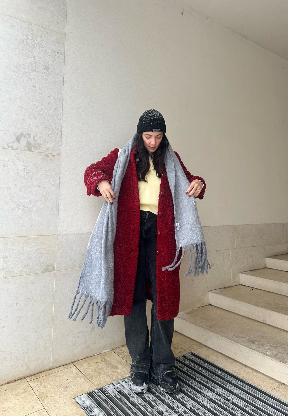 Mantello rosso texture con bottoni, maglione giallo, sciarpa grigia con frange, jeans neri e stivali, su una parete di pietra chiara e scale.
