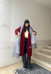 Mantello rosso texture con bottoni, maglione giallo, sciarpa grigia con frange, jeans neri e stivali, su una parete di pietra chiara e scale.