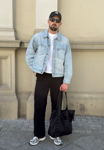 Hombre con gafas de sol, gorra negra, chaqueta vaquera azul, camisa blanca, pantalones negros y zapatillas, sosteniendo una bolsa de mano negra, de pie al aire libre.