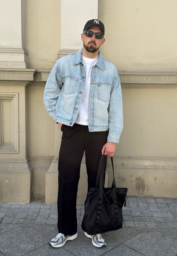 Bărbat purtând ochelari de soare, șapcă neagră, jachetă de blugi albastră, cămașă albă, pantaloni negri și adidași, ținând o geantă de umăr neagră, stând în aer liber.