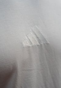 Hvid bomulds t-shirt med et hævet, sølvfarvet tre-stripes logo på forsiden. Korte fibre skaber en glat tekstur.