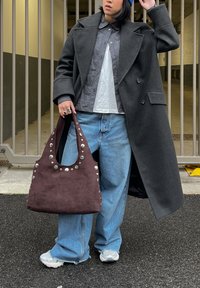 Personne portant un manteau gris foncé, une chemise à carreaux, un jean bleu, des baskets blanches, un bonnet bleu, tenant un sac à main marron clouté, debout en plein air.