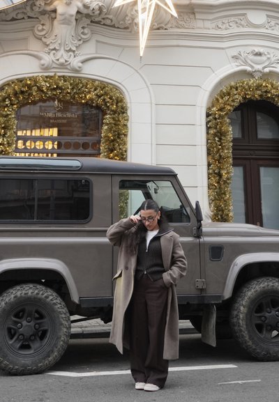 SUV vintage gris estacionado a lo largo de una calle; adornado con decoraciones doradas navideñas; una persona con abrigo y pantalones marrones está cerca.