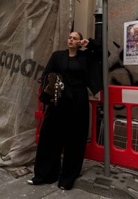 Black oversized blazer and wide-leg trousers, paired with a black top. Holding a brown bag with metal details. The urban backdrop features construction barriers.