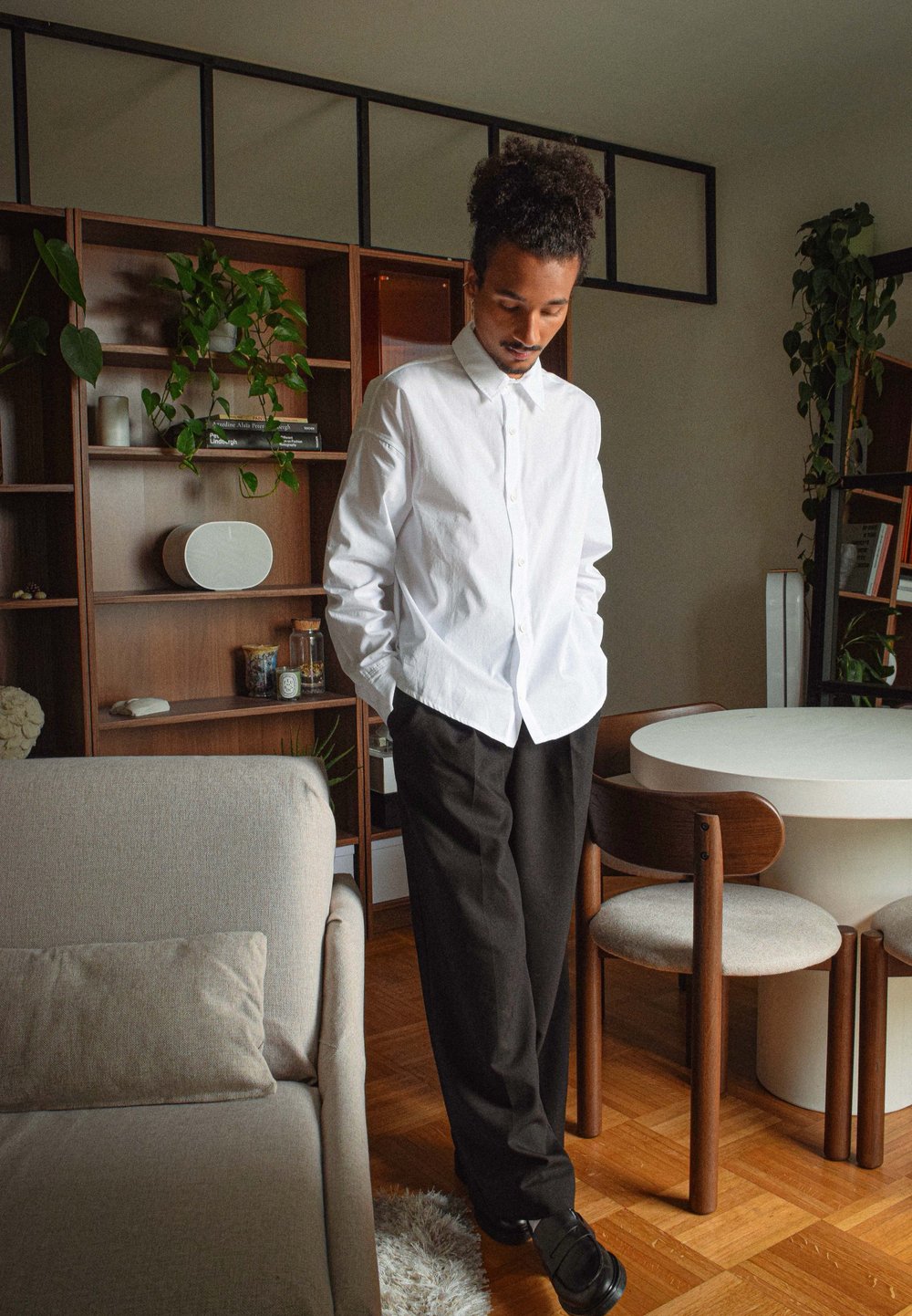 Vit skjorta med långa ärmar, kombinerad med svarta byxor. Rummet har träreglar, växter och ett runt vitt bord.