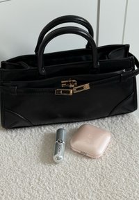 Sac à main en cuir noir avec fermoir doré reposant sur une surface blanche texturée, accompagné d'un rouge à lèvres argenté et d'un poudrier beige de KIKO.