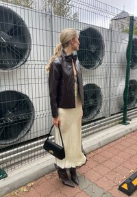 Bruine leren jas, crèmekleurige satijnen jurk met kantafwerking, zwarte handtas en puntige bruine hoge hakken. Staat bij grote ventilatoren.