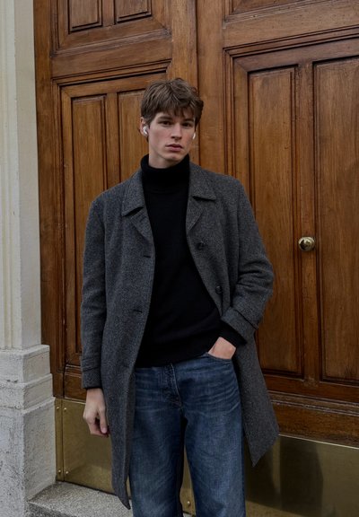 Abrigo gris de lana con doble botonadura sobre un jersey de cuello alto negro, combinado con vaqueros de mezclilla azul. Textura suave, botones prominentes, diseño clásico.