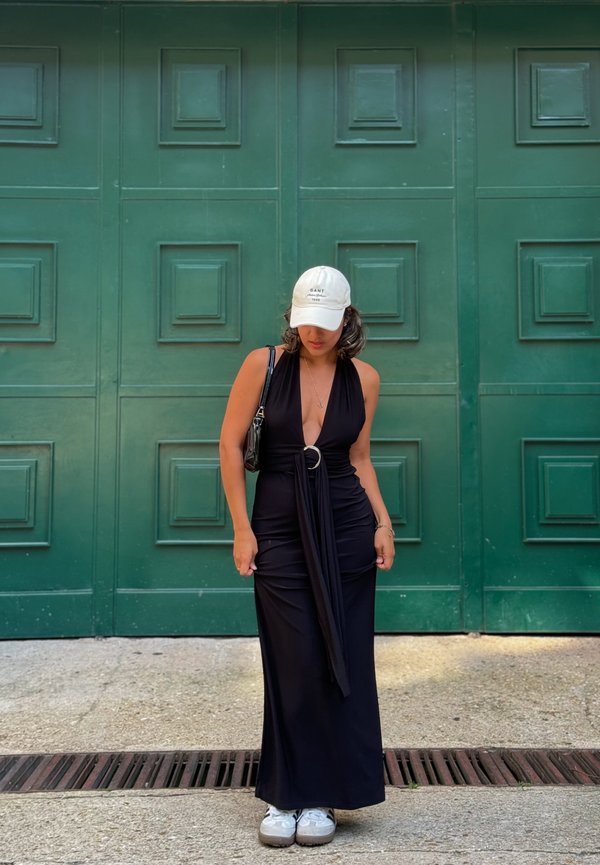 Robe noire à col halter avec un détail de nœud à l'avant, mettant en valeur un accent circulaire à la taille. Associée à des baskets blanches et une casquette légère. Fond vert.
