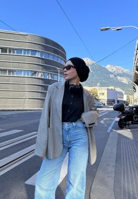 Blazer oversize gris clair, chemise noire à boutons, jeans évasés bleu clair, casque noir, lunettes de soleil, arrière-plan urbain avec des montagnes.