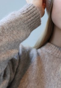 Gaiši bēšs adīts džemperis ar ribotiem aproču galiem un mīkstu, pūkainu tekstūru. Apģērbs daļēji sedz roku un plecu uz gaiša fona.