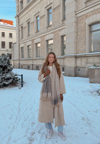 Νεαρή γυναίκα που φοράει μπεζ παλτό, κασκόλ, γάντια, τζιν και αθλητικά παπούτσια, στέκεται σε χιονισμένο έδαφος κοντά σε ένα πολυώροφο κτήριο από τούβλα.