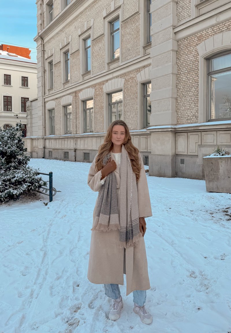 Jeune femme portant un manteau beige, une écharpe, des gants, un jean et des baskets, debout sur un sol couvert de neige près d'un bâtiment en briques à plusieurs étages.