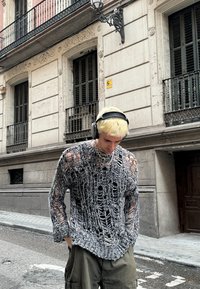 Zwart-witte gebreide trui met een open weefdesign en versleten details, gedragen met olijfgroene cargo-broek en koptelefoon.