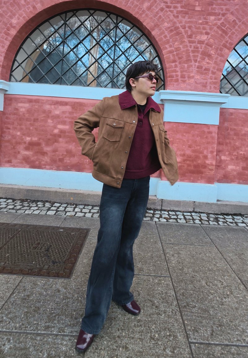 Personne portant des lunettes de soleil, une veste marron, un pull bordeaux et un jean foncé, debout sur un trottoir en pierre devant un mur de briques roses avec des fenêtres en arc.