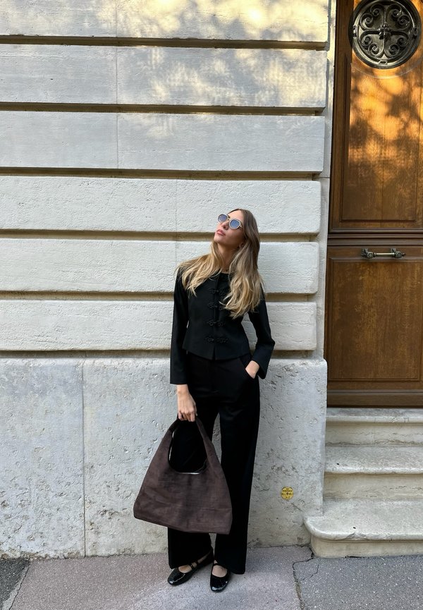 Mujer con el pelo largo y rubio, vestida de negro y con gafas de sol, sosteniendo una gran bolsa marrón, de pie junto a una pared de piedra blanca cerca de una puerta de madera.