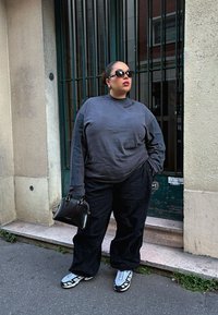 Grå langermet skjorte med høy krage, svarte vide bukser og grå joggesko med hvite detaljer; holder en svart håndveske.