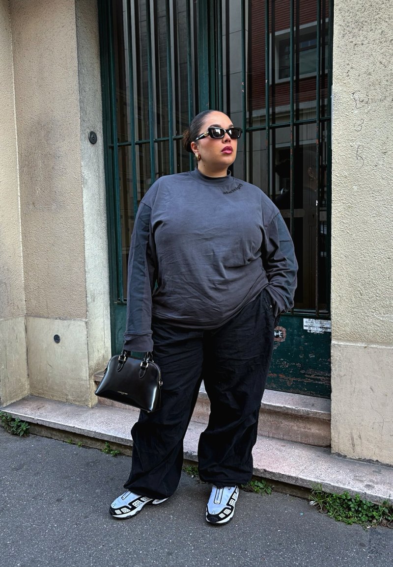 Grijze lange mouwen shirt met een hoge kraag, zwarte losse broek en grijze sneakers met witte accenten; houdt een zwarte handtas vast.