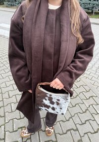 Bruine overjas met lange mouwen, gecombineerd met een sjaal. Houdt een gespikkelde bruin-witte handtas vast en draagt bruine schoenen met patroon.