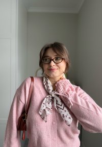 Jeune femme portant des lunettes, un cardigan rose et une écharpe à motifs, debout à l'intérieur avec un sac à bandoulière marron.