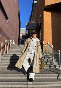 Personne en manteau long beige, sweat à capuche gris et pantalon de survêtement descendant des escaliers en pierre extérieurs entre des bâtiments en briques, tenant un sac vert olive.