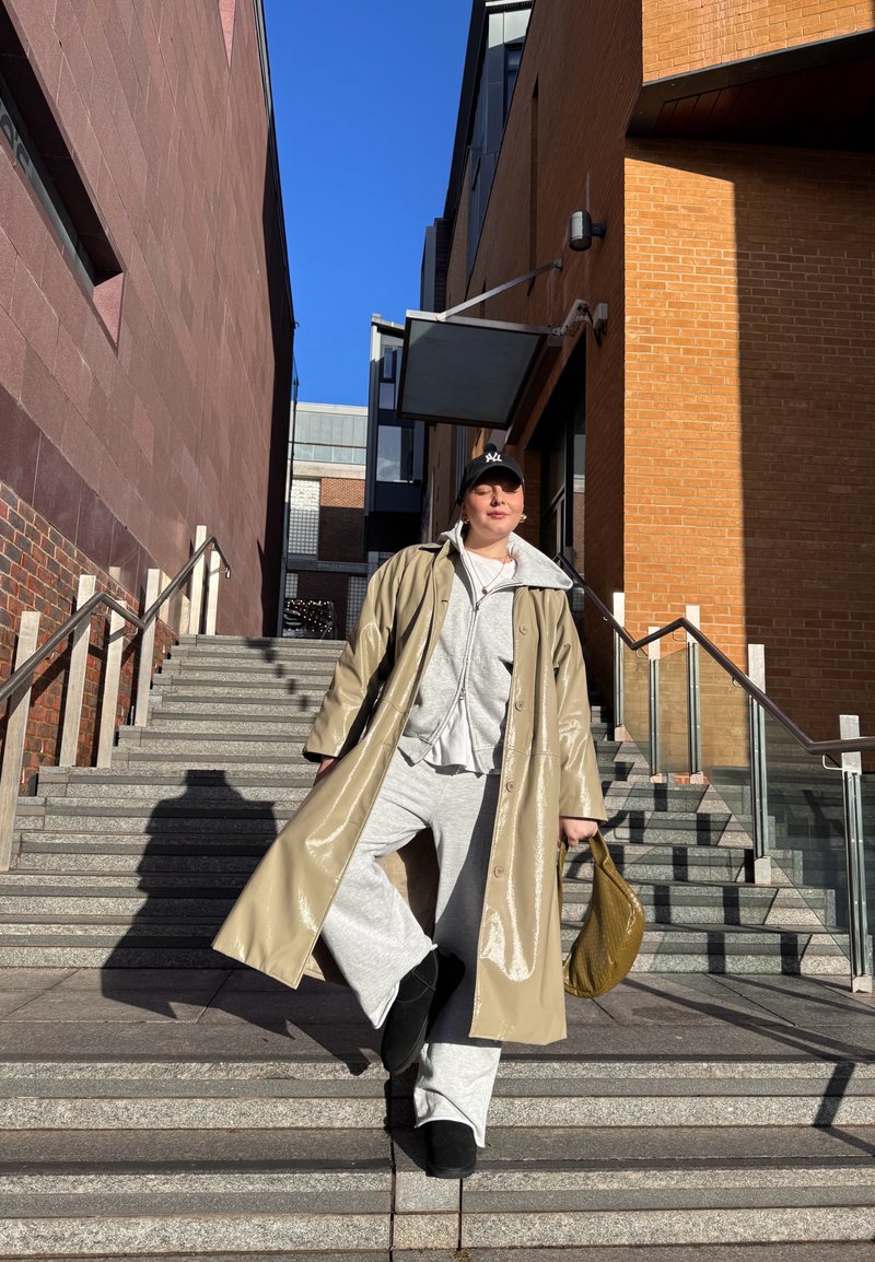 Personne en manteau long beige, sweat à capuche gris et pantalon de survêtement descendant des escaliers en pierre extérieurs entre des bâtiments en briques, tenant un sac vert olive.