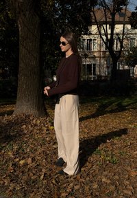 En person iført en mørkebrun sweater, beige bukser med vide ben og sorte sko står blandt faldne blade med et træ og bygninger i baggrunden.