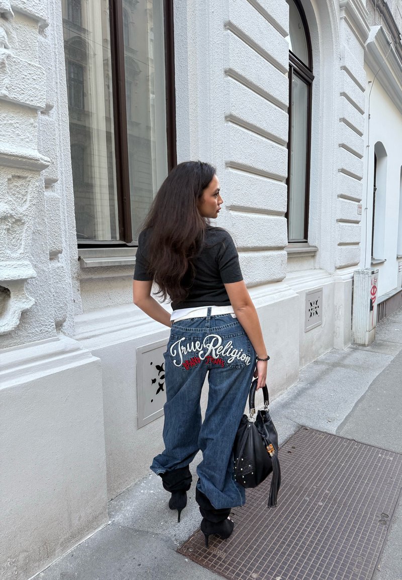 Femme aux longs cheveux foncés portant un top noir et un jean large avec le texte « True Religion », tenant un sac à main noir, debout dans une rue européenne.