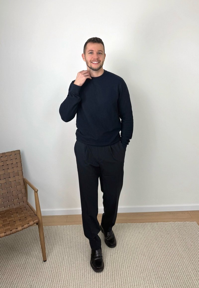 Smiling man in black sweater and pants standing on a rug beside a woven wooden chair against a plain white wall.
