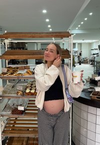 Έγκυος άτομο με μαύρη μπλούζα και λευκή oversized ζακέτα, κρατώντας ένα φλιτζάνι καφέ, στέκεται δίπλα σε ξύλινα ράφια με αρτοσκευάσματα.