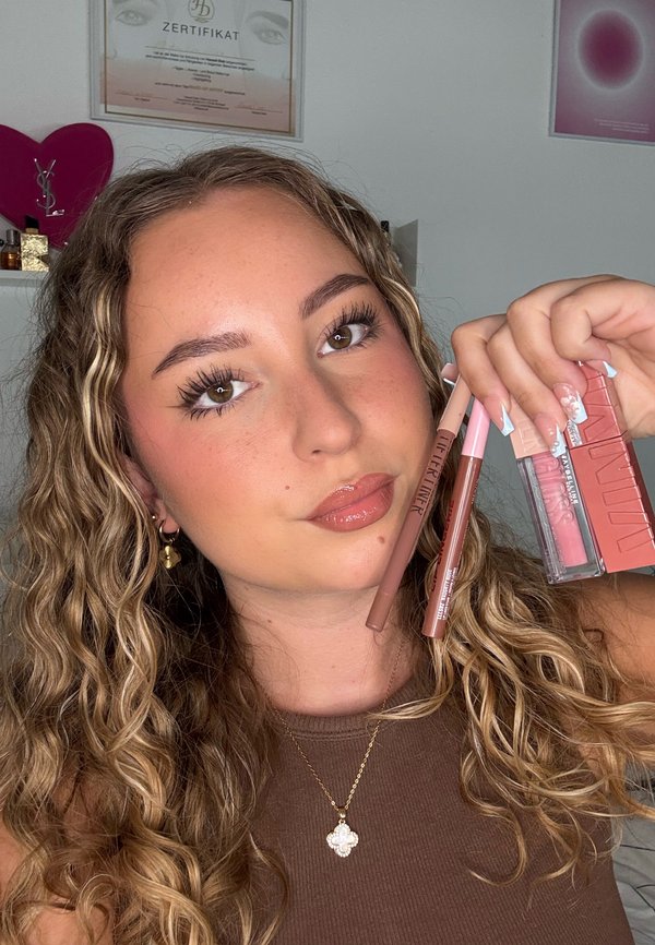 Jeune femme aux cheveux bouclés tenant plusieurs produits de maquillage des lèvres près de son visage, portant un haut marron et un collier avec pendentif fleur.