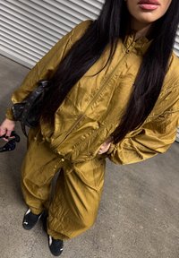 Złotobrązowy dres z zamkiem błyskawicznym i elastycznymi mankietami, wykonany z teksturowanej tkaniny. Noszony z czarnymi trampkami i małą torbą.