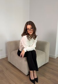Jeune femme aux cheveux foncés et portant des lunettes assise sur un fauteuil beige, vêtue d'un pull blanc, d'un pantalon noir et de talons noirs dans une pièce minimaliste.