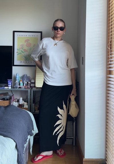 Persona que lleva gafas de sol negras, una camisa blanca de gran tamaño, una falda negra con un diseño de sol beige, chanclas rojas, sosteniendo un bolso beige, de pie en interiores.